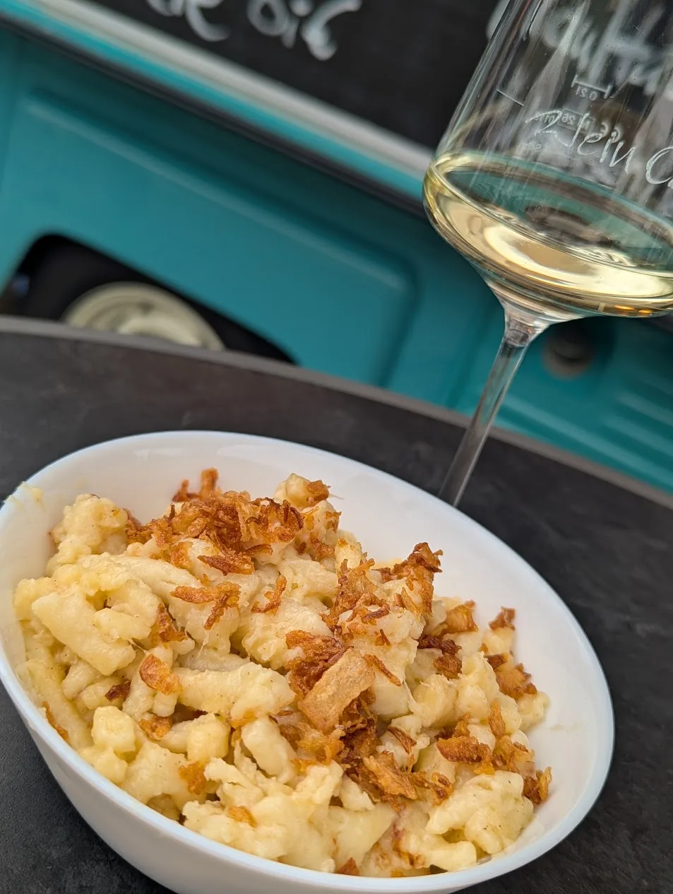 Bergkäsespätzle mit Röstzwiebeln, dazu ein Glas Weißwein vor dem Foodtruck